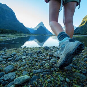 Zwei Beine einer Person in Wanderschuhen an einem See vor Bergpanorama.