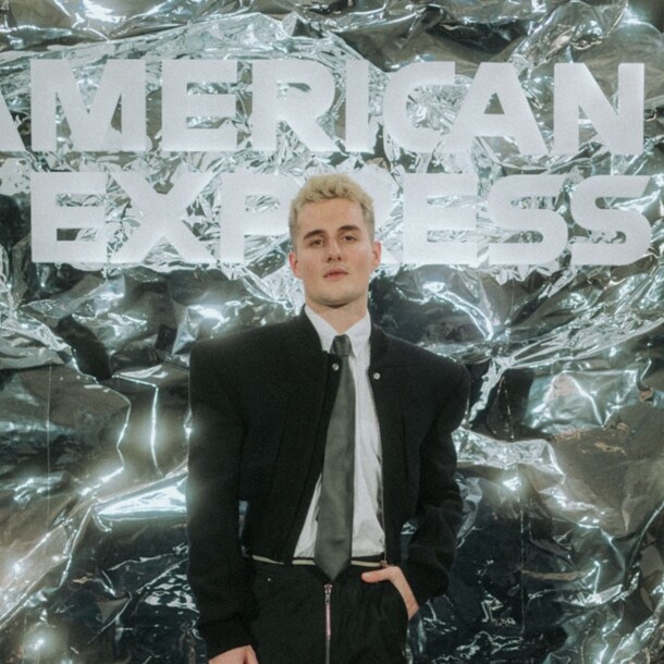 Luis Freitag von den Elevator Boys im schwarzem Anzug mit weißem Hemd und Krawatte vor einer silbernen Wand mit weißem American Express Schriftzug.