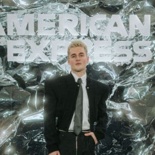 Luis Freitag von den Elevator Boys im schwarzem Anzug mit weißem Hemd und Krawatte vor einer silbernen Wand mit weißem American Express Schriftzug.