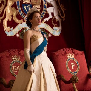 Eine junge Königin mit Krone und blauer Schärpe über einem beigefarbenen Kleid vor zwei roten Polsterthronen an einer roten, samtverkleideten Wand mit Wappen.