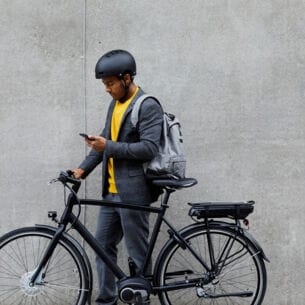 Ein junger Mann mit Rucksack und Fahrradhelm steht neben seinem E-Bike und schaut auf sein Smartphone.
