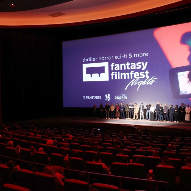 Eine Personengruppe steht vor einer Leinwand auf einer Bühne vor Publikum in roten Samtsesseln in einem Kinosaal.