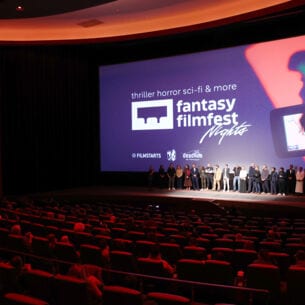 Eine Personengruppe steht vor einer Leinwand auf einer Bühne vor Publikum in roten Samtsesseln in einem Kinosaal.
