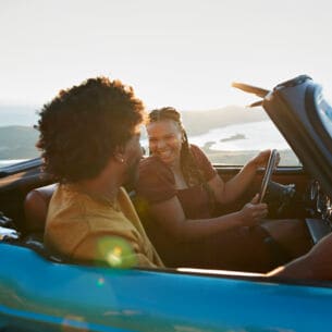 Ein junges Paar in einem blauen Cabriolet schaut sich fröhlich an, im Hintergrund eine Küstenlandschaft.