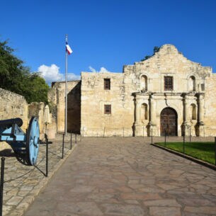 Frontfassade der Kirche einer spanischen Missionsstätte, davor ein Platz mit Texanischer Flagge und einer Kanone.