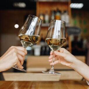Nahaufnahme von Händen und Unterarmen von zwei Personen, die jeweils ein gefülltes Weinglas halten und anstoßen.