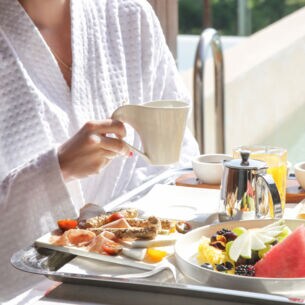 Eine Person sitzt an einem Tisch und hält eine Tasse in der Hand. Vor ihr steht ein Frühstückstablett mit Essen und Getränken.
