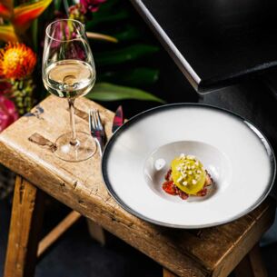 Ein weißer Teller mit einer kreativ angerichteten Vorspeise steht neben einem Glas Weißwein auf einem Holzschemel, daneben ein Blumenbouquet.