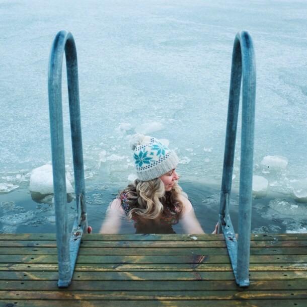 Eine Frau mit Wollmütze hält sich beim Baden in einem zugefrorenen See an dem Handlauf einer Treppe an einem Holzsteg fest.
