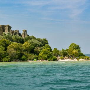Eine antike Ruine, umgeben von üppiger Natur, an einem Sandstrand mit Personen am türkisblauen Wasser.