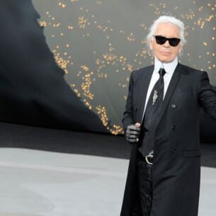 Karl Lagerfeld mit Sonnenbrille im schwarzen Jackett winkt vom Laufsteg einer Modenschau.