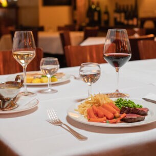 Zwei auf weißen Tellern angerichtete Speisen stehen neben Weingläsern auf einem Tisch mit weißer Stofftischdecke in einer Brasserie.