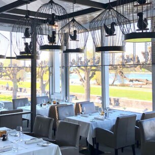 Speiseraum des Restaurants OX Royal mit eingedeckten Tischen und dekorativen Vogelkäfigen an der Decke in einem Wintergarten mit Panoramablick an der Uferpromenade eines Flusses.