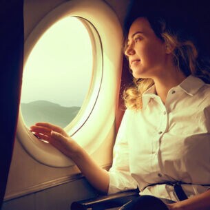 Eine junge Frau in weißer Bluse schaut aus dem Fenster eines Flugzeuges auf eine Landschaft.