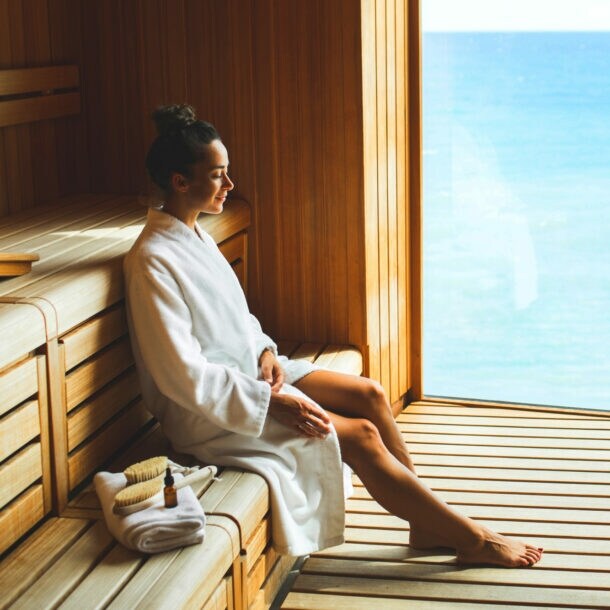 Eine junge Frau im weißen Bademantel sitzt entspannt in einer Holzsauna mit Panoramafenster am Meer.