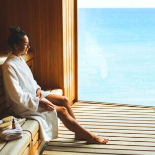 Eine junge Frau im weißen Bademantel sitzt entspannt in einer Holzsauna mit Panoramafenster am Meer.