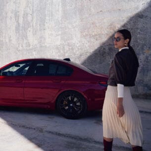 Eine elegante Frau mit Sonnenbrille vor einem roten BMW vor einer Betonwand.