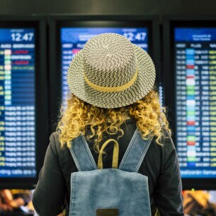Rückansicht einer Frau mit Strohhut vor einer Anzeigetafel am Flughafen.