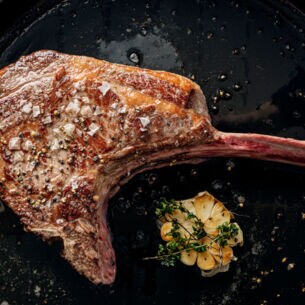 Ein scharf angebratenes Tomahawk-Steak in einer Pfanne. Daneben liegt eine aufgeschnittene Knoblauchknolle, die mit Thymian getoppt ist.