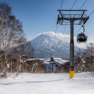 Skilift und Schneelandschaft mit Bäumen. Im Hintergrund ein schneebedeckter Berg und blauer Himmel.