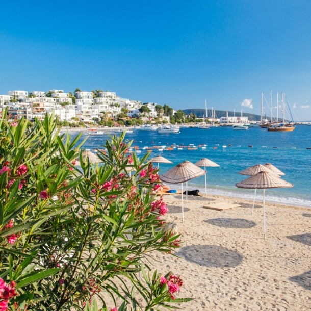 Strand in Bodrum mit Sonnenschirmen mit weißen Häusern an der Küste im Hintergrund.