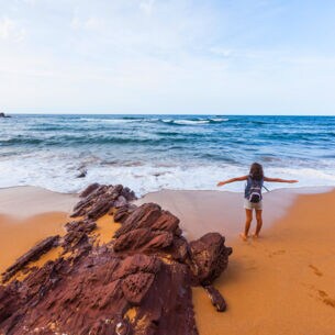 Eine Person steht mit dem Rücken zur Kamera und neben einem Felsen an einem Strand und schaut mit ausgebreiteten Armen aufs Meer