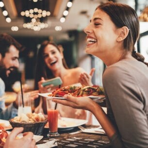 Eine Gruppe fröhlicher Personen in einem Restaurant. Eine lachende Frau hält einen Teller in der Hand.