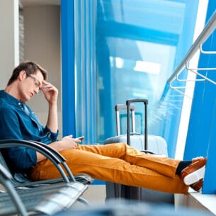 Ein Mann, der am Flughafen auf einer Bankreihe mit ausgestreckten Beinen sitzt. In einer Hand hält er ein Smartphone, die andere Hand ist an seiner Schläfe. Neben ihm steht ein Koffer.