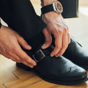 Männerhände schließen den seitlichen Schnallriemen eines schwarzen Monkstrap-Schuhs mit Lochmusternaht auf der Zehenkappe.
