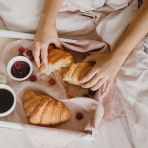 Auf einem Bett liegt ein Tablett mit Croissants, einer Tasse Kaffee, ein Schälchen Marmelade, einem Löffel und Früchten. Dahinter sitzt eine unkenntliche Person, die eines der Croissants auseinanderreißt.