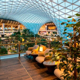 Moderne Lounge auf einer Empore in einem begrünten Flughafenterminal mit Bäumen unter einer geschwungenen Glaskuppel.