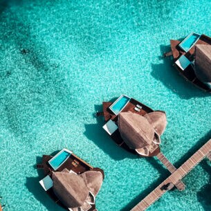 Eine Luftaufnahme einer Hotelanlage mit privaten Suiten samt Pool auf den Malediven, die in einer Linie an einem Steg angelegt und von türkisblauem Wasser umgeben sind