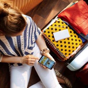 Aufsicht einer Frau, die neben einem gepackten Koffer auf dem Boden sitzt und auf ihrem Smartphone eine Reise bucht.