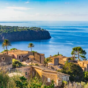 Ein altes Dorf an der Küste Mallorcas. Im Hintergrund eine grün überwachsene Landzunge, die in das Meer hineinragt.