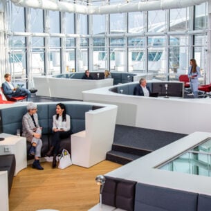 Mehrere Personen sitzen in einer modernen Sitzlandschaft einer Flughafenlounge mit Panoramafenstern.