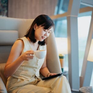 Eine Geschäftsfrau sitzt mit einer Tasse Kaffee in einem Sessel in einer Flughafenlounge und schaut auf ein Tablet in ihrer Hand.