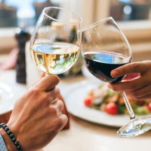 Zwei Personen, die jeweils ein Glas Wein halten und anstoßen. Auf dem Tisch vor ihnen stehen Teller mit Speisen.