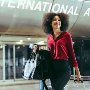 Eine elegante, lächelnde Frau mit Handgepäck vor einem Ausgang am Flughafen.