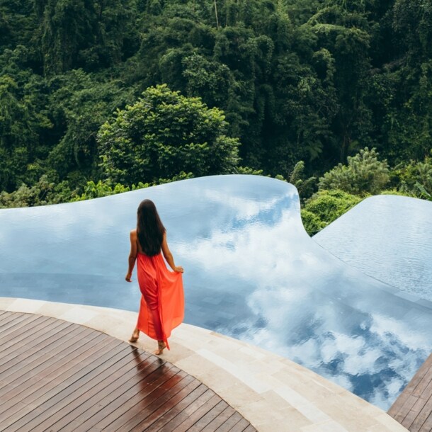 Rückansicht einer Frau im langen Sommerkleid an einem Infinitypool auf einer Terrasse, umgeben von tropischem Regenwald.