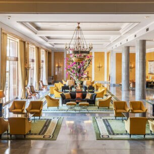 Ein elegantes Hotelfoyer mit Marmorsäulen und gelben Sesseln, in der Mitte ein großes Blumenbouquet.