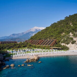Ein Hotelresort in einer mediterranen Bucht mit Sandstrand, im Hintergrund eine Berglandschaft.