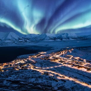 Beleuchtete Ortschaft in schneebedeckter Fjordlandschaft unter Polarlichtern bei Nacht.