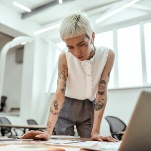 Eine junge tätowierte Frau steht am Schreibtisch und blickt auf verschiedene Entwürfe.