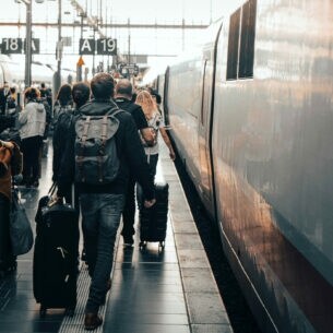 Reisende mit Gepäck laufen am Gleis in einem großen Bahnhof.