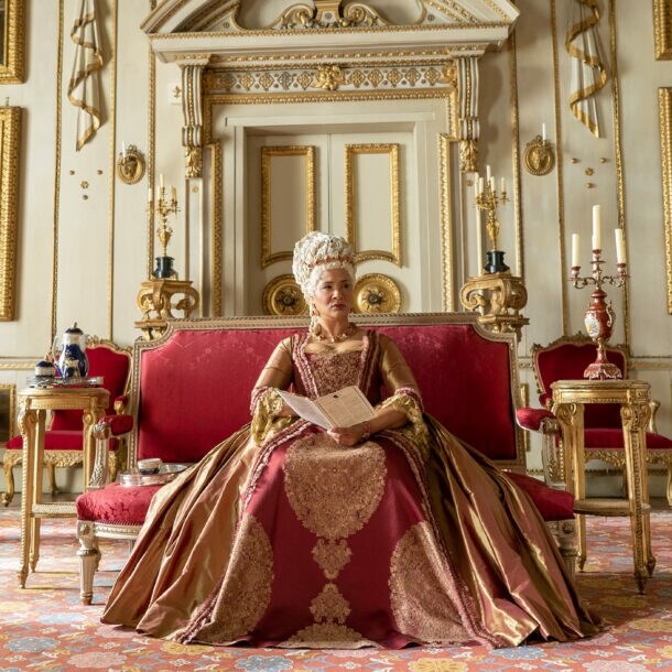 Eine Königin sitzt auf einem roten Samtsofa in einem Palast mit weiß-goldenem Dekor.