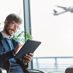Ein Mann, der am Flughafen sitzt und ein Tablet bedient. Vor ihm steht ein Koffer und im Hintergrund startet ein Flugzeug.
