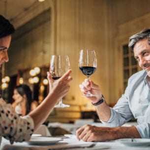 Eine Frau und ein Mann sitzen in einem Restaurant am Tisch, heben je ein Weinglas hoch und lächeln sich an.