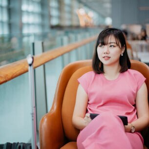 Eine asiatische Frau in einem pinken Kleid sitzt in einem braunen Ledersessel auf einer Galerie einer Flughafenlounge.