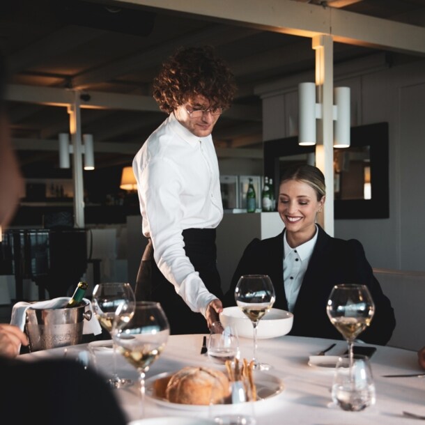 Eine lächelnde Frau wird in einem Restaurant von einem Kellner bedient, der einen Teller vor ihr auf den weiß eingedeckten Tisch stellt.