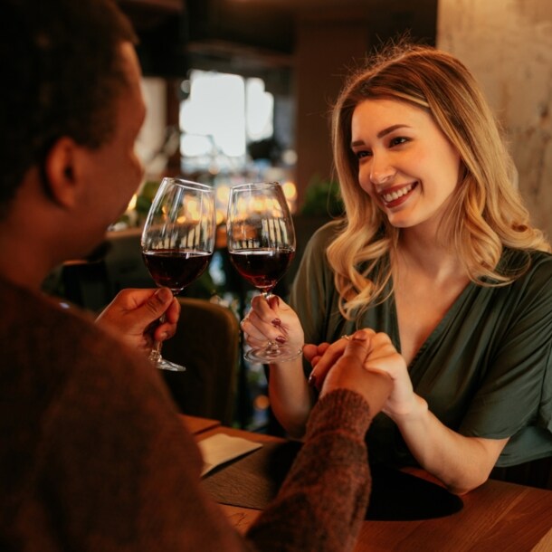 Eine lächelnde Frau sitzt einem Mann an einem Restauranttisch gegenüber und hält seine Hand. In der anderen Hand halten sie je ein Glas Rotwein.